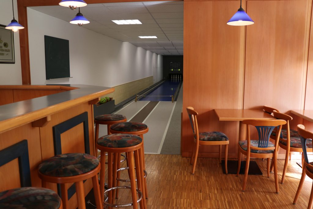 Das Bild zeigt eine Bar mit Barhockern, daneben Tische mit gepolsterten Stühlen und dahinter eine Kegelbahn.